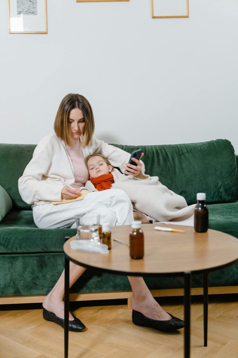 A mother comforts her sick child on a sofa while holding a smartphone, showcasing family care.